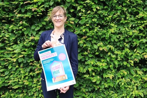 Bürgermeisterin Ute Dülfer hält ein Plakat zum Heimat-Preis 2022 in der Hand. Sie freut sich auf Vorschläge für den Heimatpreis.