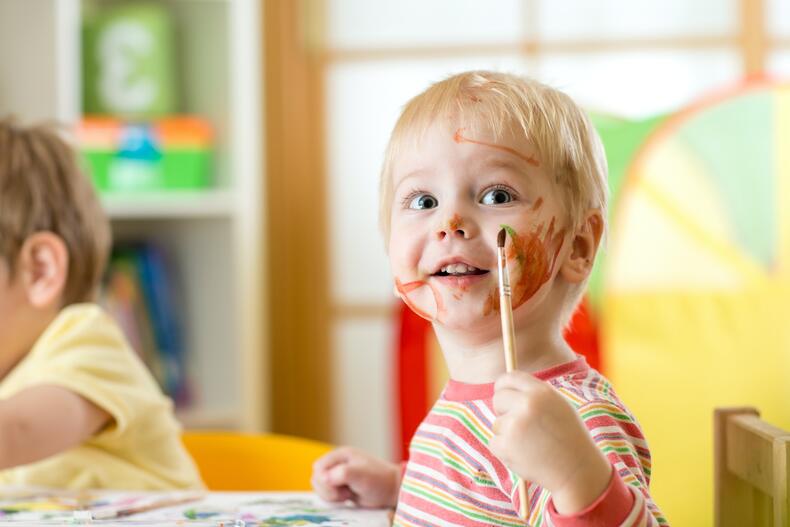 Kleiner Junge in der Kindertageseinrichtung, welcher sich mit einem Pinsel im Gesicht malt.